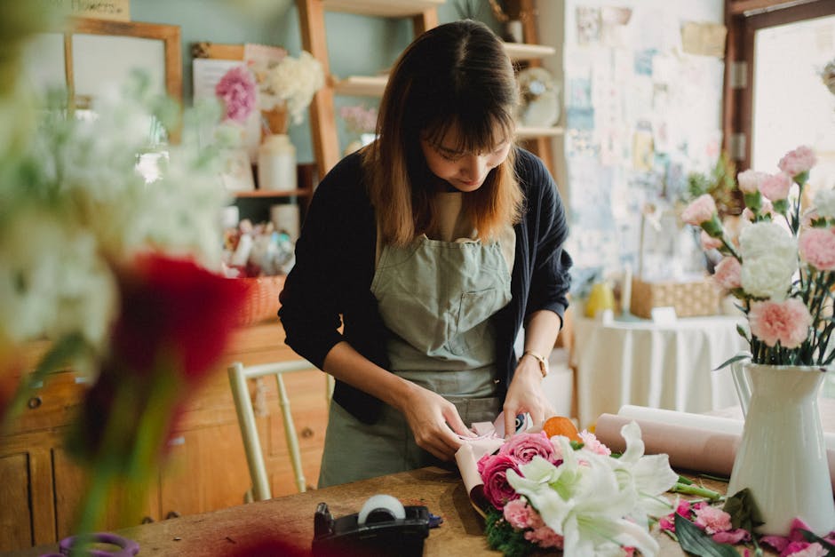 A florist with shoulder-length brown hair and bangs, wearing a beige apron over a black top, is carefully arranging a bouquet of fresh pink roses, white lilies, and other colorful flowers on a wooden