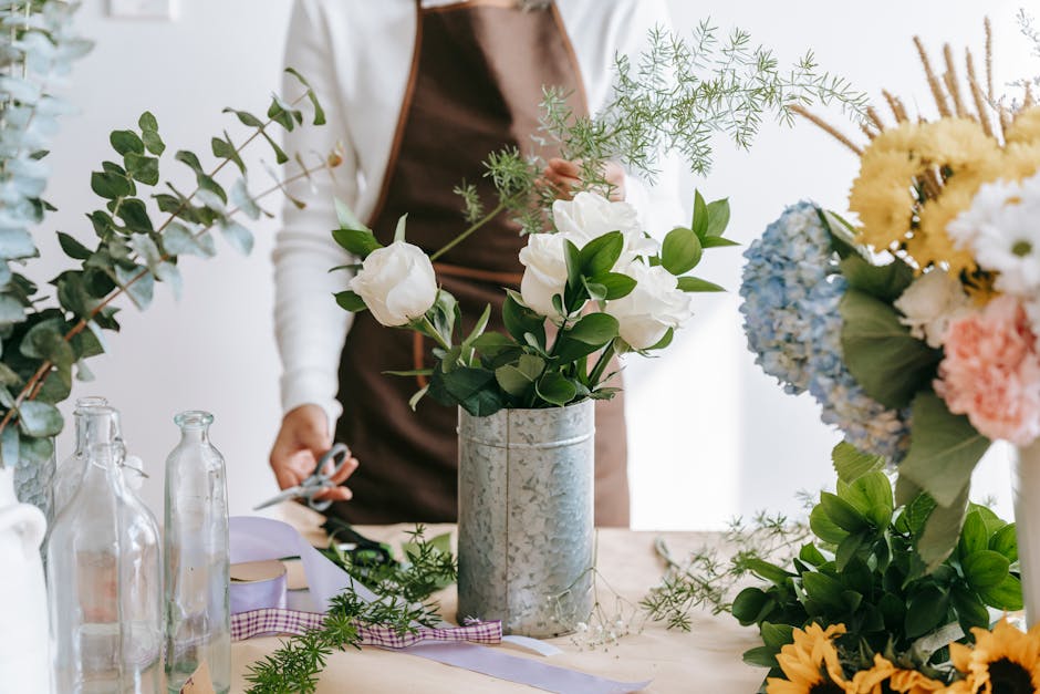 A florist wearing a brown apron and white shirt arranging a floral bouquet with white roses and green foliage in a galvanized metal vase. The table is covered with various flower arrangements, includi
