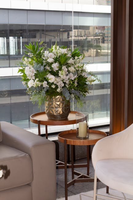 A large, elegant floral arrangement featuring white flowers such as lilies and roses, complemented by lush green foliage, is displayed in a textured, dark-colored vase on a round wooden table near a w