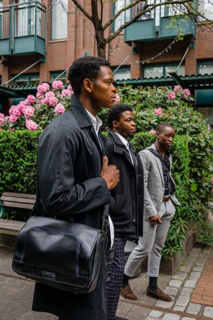 This image depicts three young men standing outdoors against a backdrop of blooming pink hydrangea bushes and a residential brick building with blue balconies. The man in the foreground has short curl