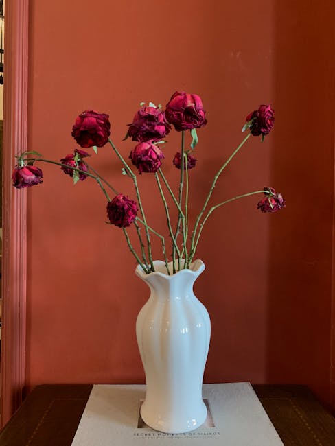 A white ceramic vase with a wavy, ribbed design holding a bouquet of wilted, deep red roses with slightly shriveled petals and green stems. The arrangement is placed on a dark wooden surface against a