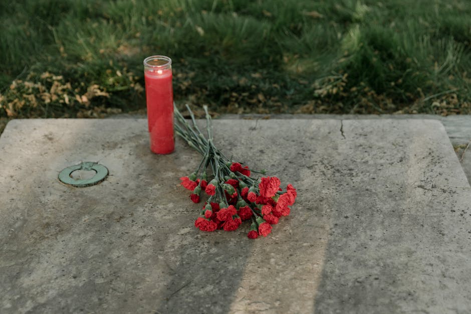 A still life scene on a weathered concrete memorial table features a red glass candle holder with a lit candle inside, and a bouquet of red carnations with green stems and leaves, lying across the sur