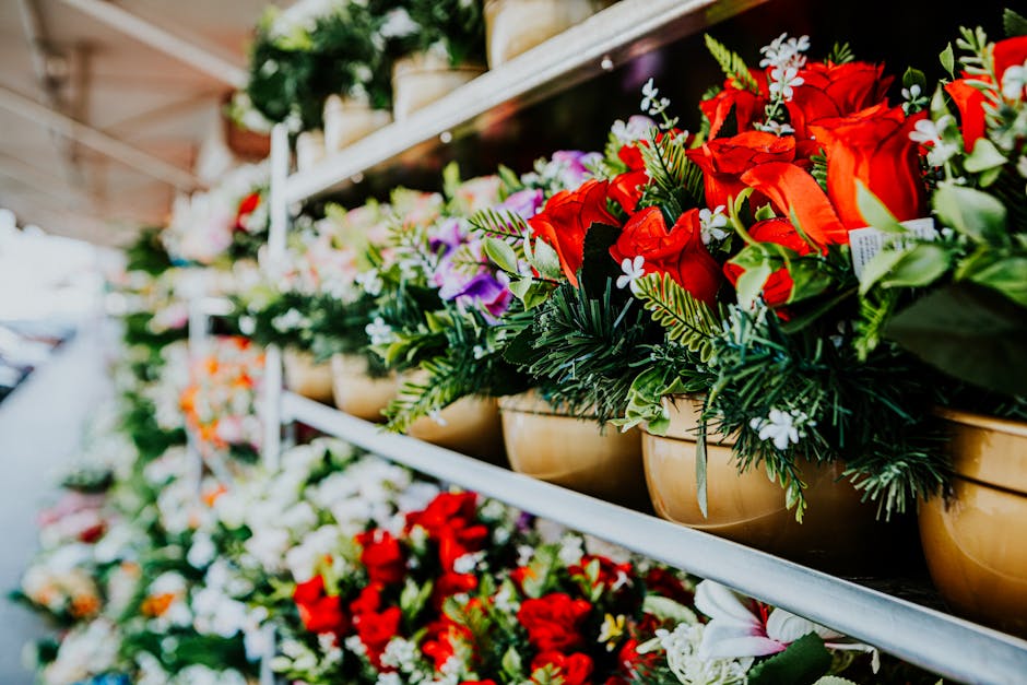 A row of vibrant floral arrangements in beige pots on display at a flower market stall, featuring red roses, white daisies, and purple lisianthus, accented with green foliage. The flowers are fresh, w
