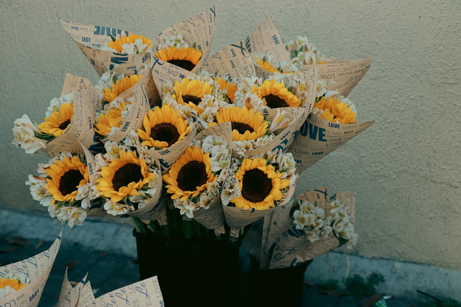 A bouquet of fresh sunflowers with bright yellow petals and dark centers, complemented by smaller white and yellow flowers, wrapped in decorative paper printed with words like 'LOVE' and 'PEACE' in bl